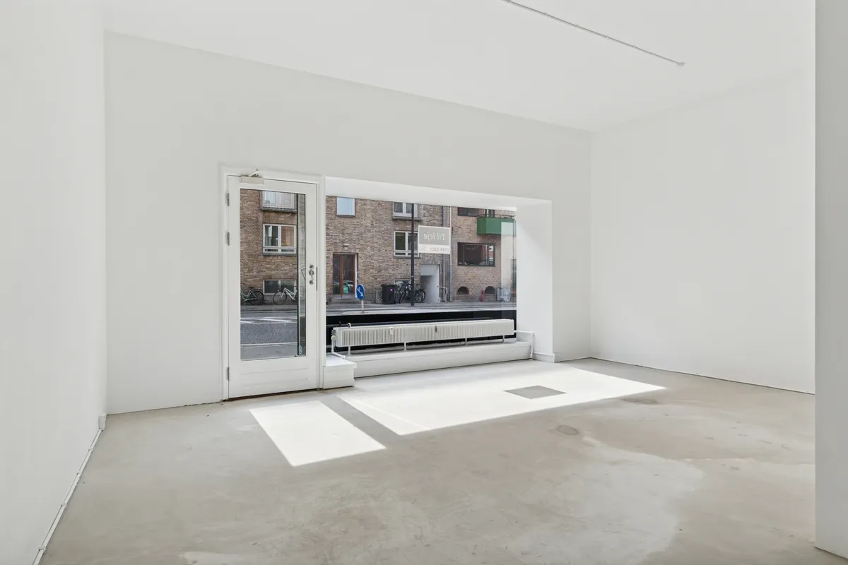 Lyst og rent butikslokale med hvide vægge og gulv og et stort glasvindu med udsigt til gaden med cykler og bygninger