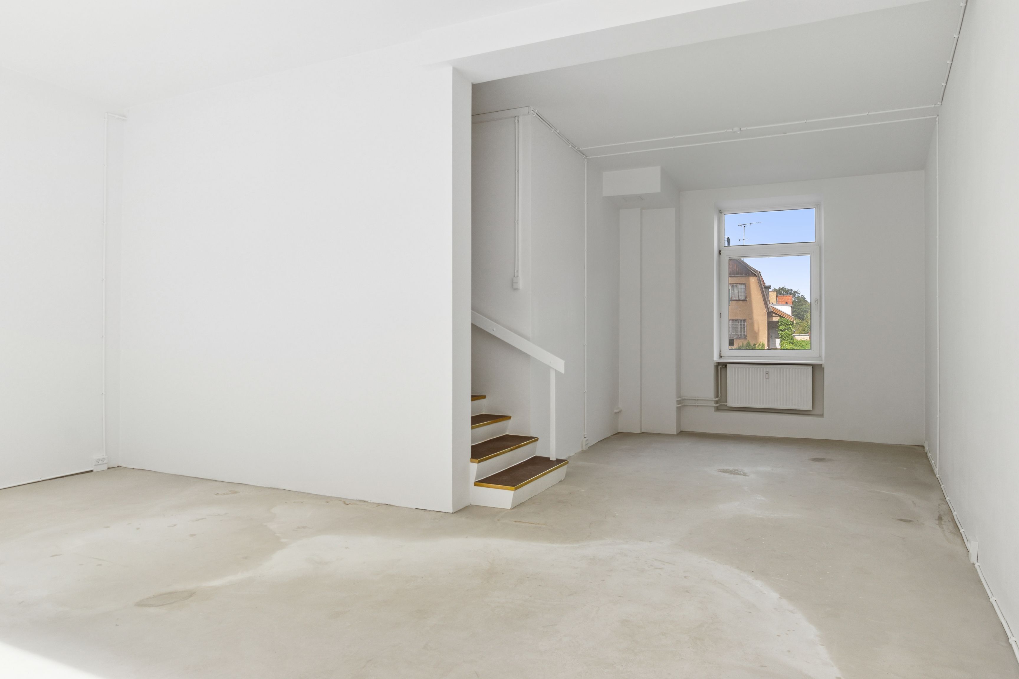 Et tomt rum med en hvid væg og gulv, stige og vinduesåbning mod en gård. Et hvidt radiatorpanel er installeret i væggen.