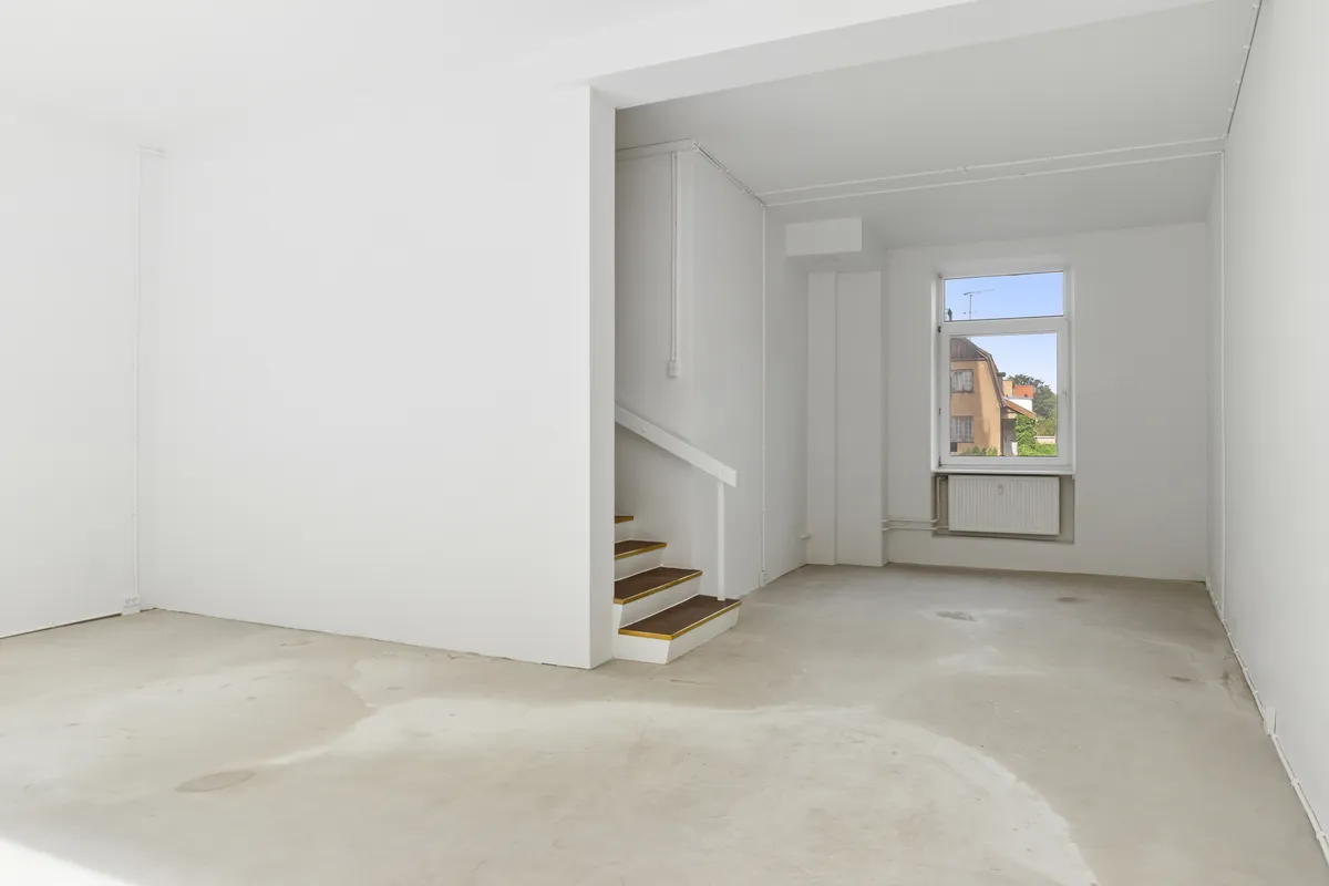 Et tomt rum med en hvid væg og gulv, stige og vinduesåbning mod en gård. Et hvidt radiatorpanel er installeret i væggen.