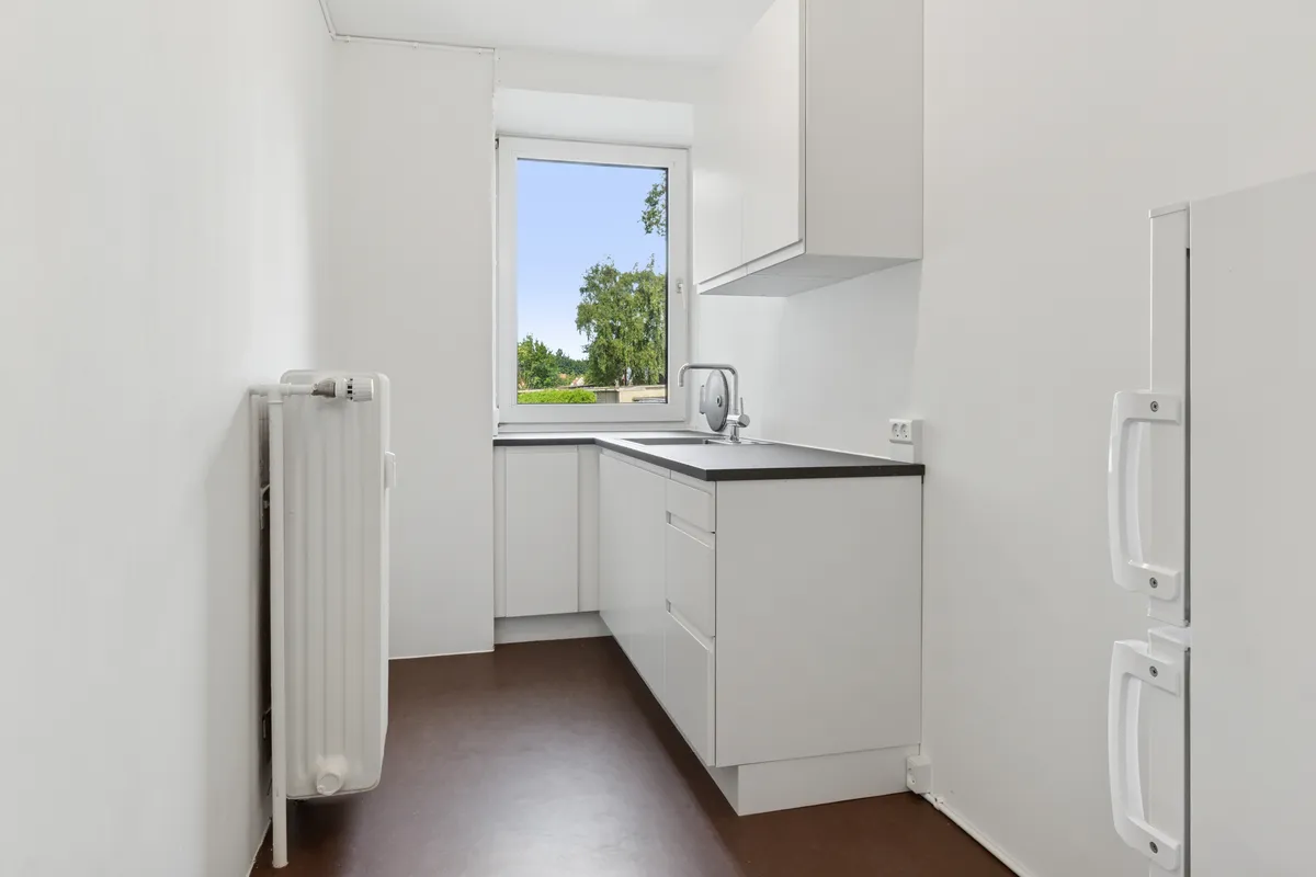 Rummeligt hvidt køkken med lyse ruder, mørkt gulv, lyseskabe, køkkenvask og lysegreb på hvidt køleskab, samt radiator i hvidt vægpanel med lysegrå gulvbelægning
