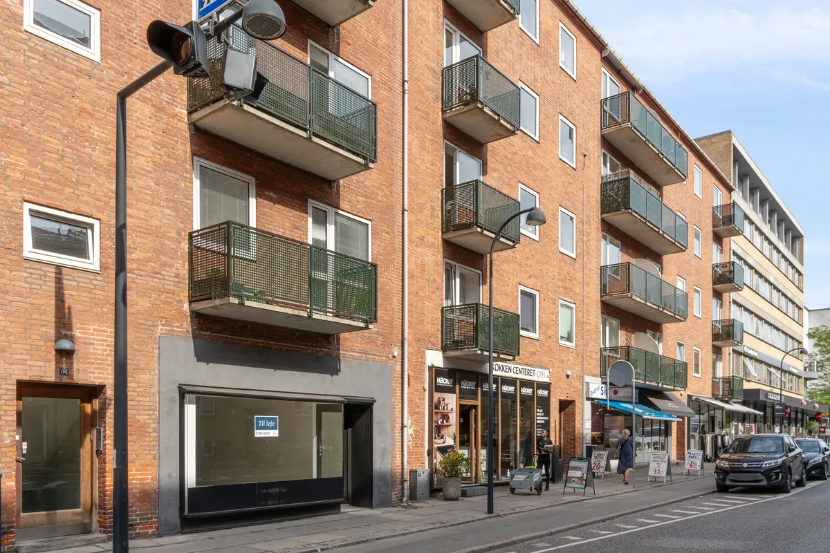 Mennesker går forbi et butikslokale på Ordrupvej 64 i Charlottenlund, der er udstyret med flere balkoner og en stor vinduesfront, og biler parkeret langs siderne af gaden