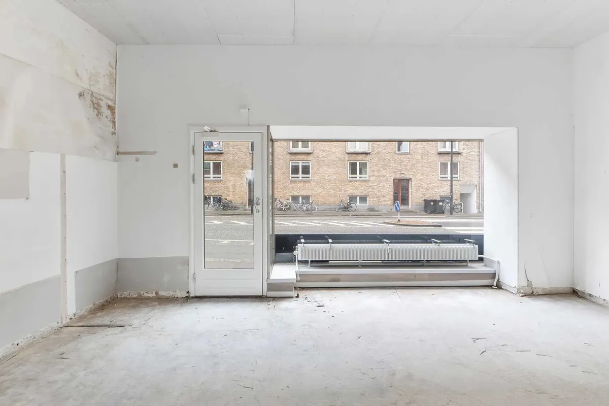 Et tomt rummeligt butikslokale med store vinduer og åbne døre mod en bygning og gade med parkerede cykler