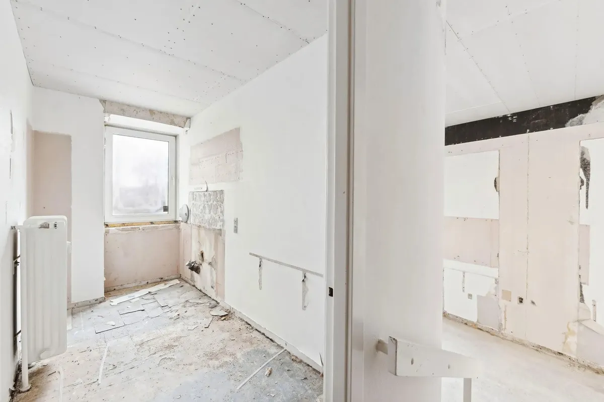 En renoveret og forladt stue med hvide vægge og loft samt en åben dør og et vindue med en hvid gardin