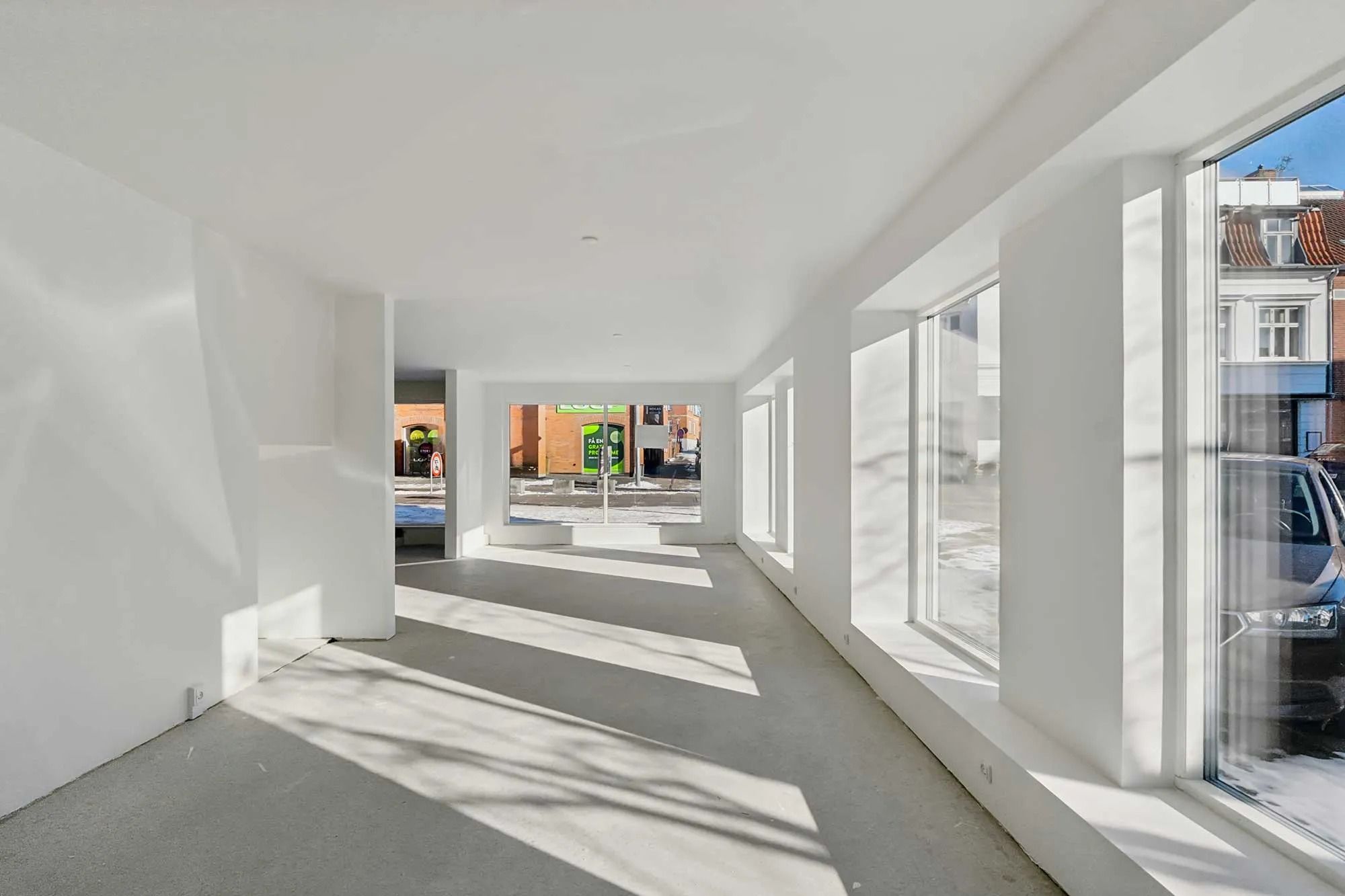 Foto af et tomt, rummeligt butikslokale med hvide vægge og gulv samt store vinduer mod gaden