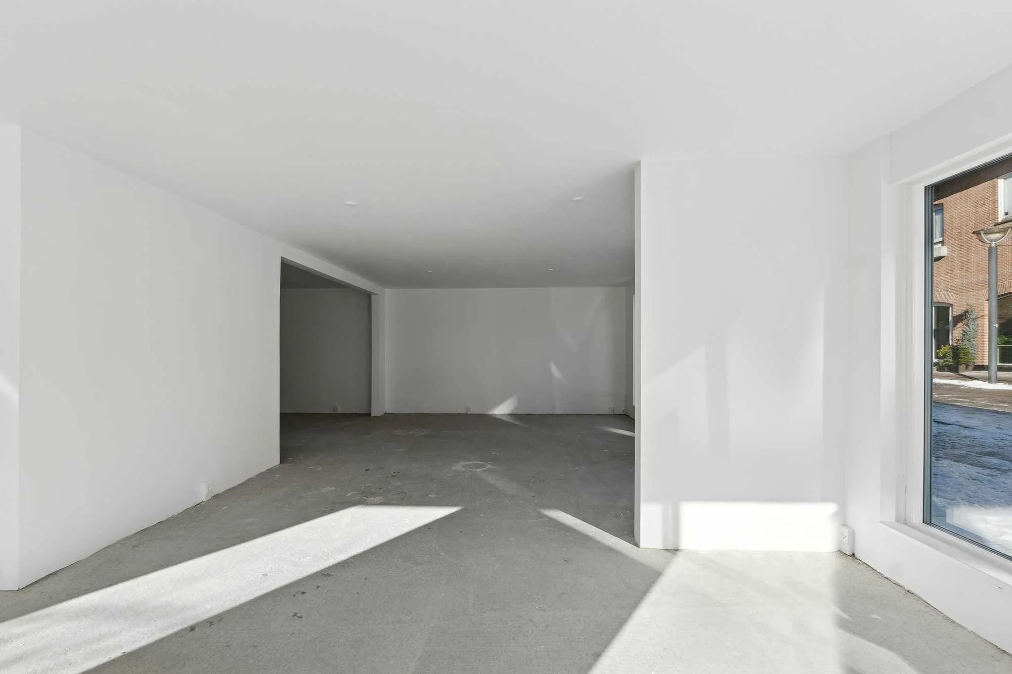 Rummeligt og ubenyttet lagerrum med hvide vægge og gulve samt en stor vinduesåbning mod en gård med sne og en lampestang