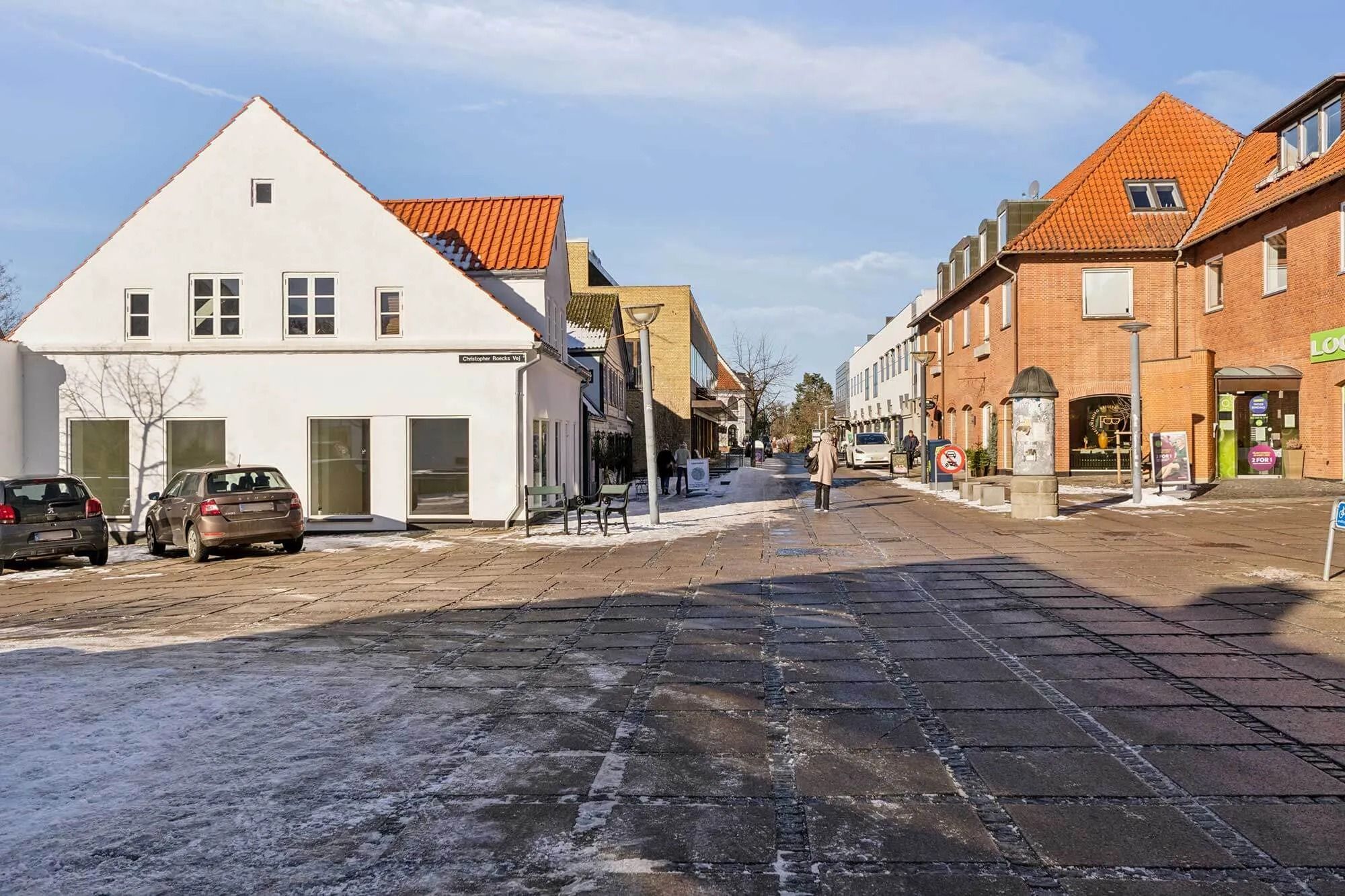 Mennesker vandrer langs en højtliggende gågade i Fredensborg, med udsigt mod et hvidt bygningshus med orange tag og flere