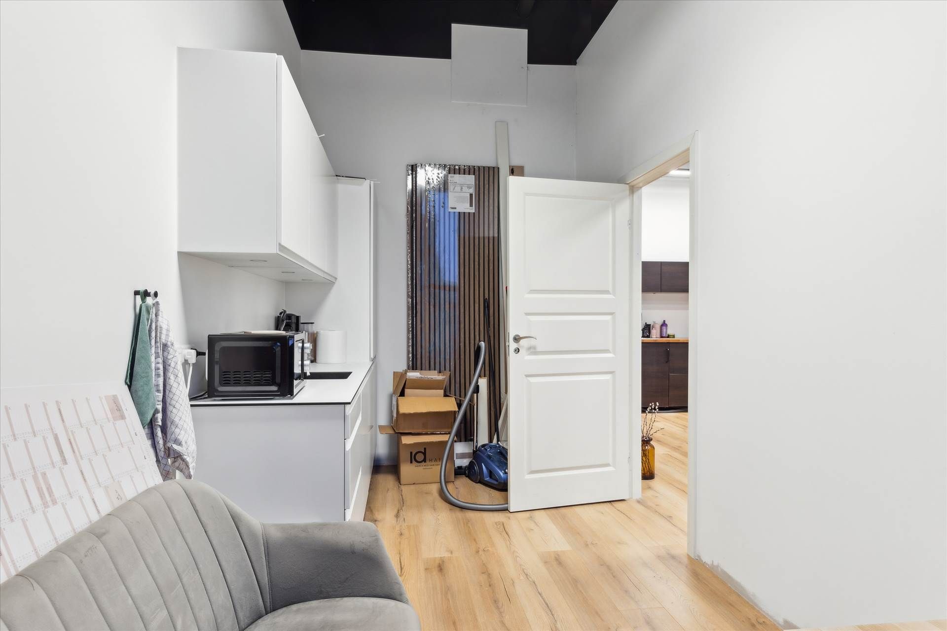 Et lyse, minimalistisk indrettet køkken med hvide vægge, trægulv og lyseskabe samt en mikrobølgeovn og et støvsuger.