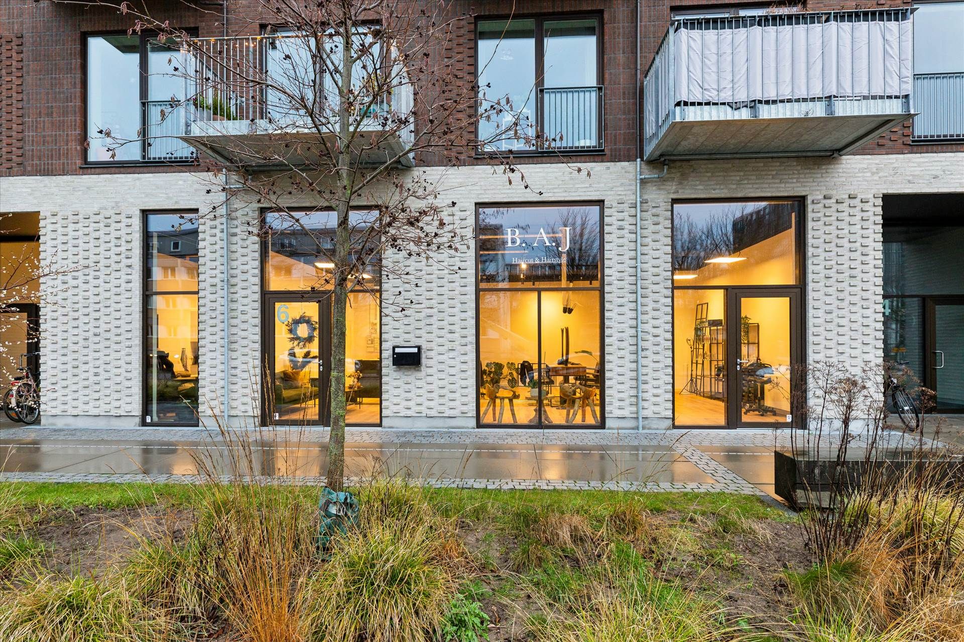 Butik med glasdøre og vinduerne med indskriften B.A.J Hair & Hairstyle, balkoner og et grønt område med græs og planter