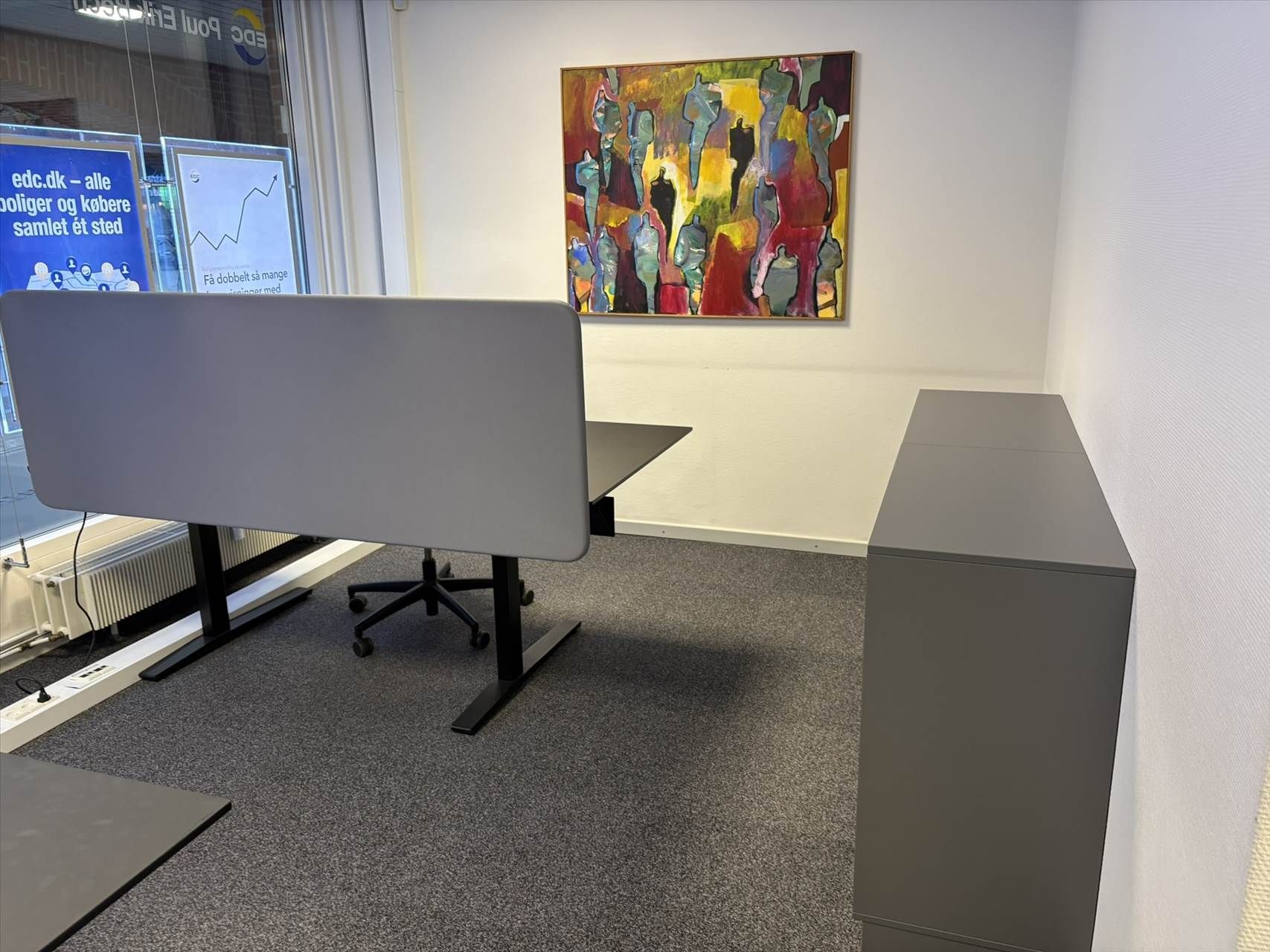 Kontor med bord og stol samt en væg med maleri og tekst. Der er også et kabinet til opbevaring og et vinduesvisker.