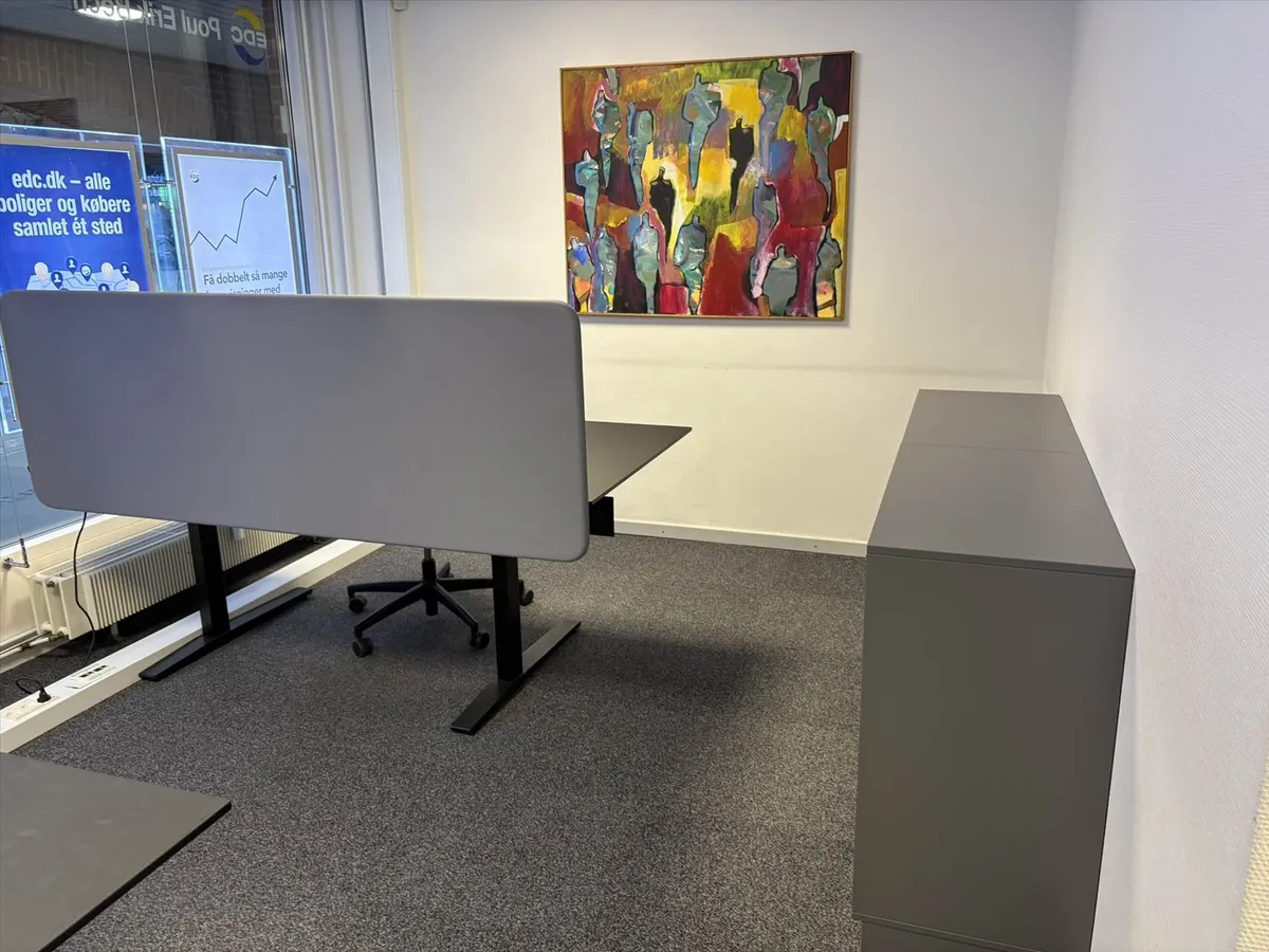 Kontor med bord og stol samt en væg med maleri og tekst. Der er også et kabinet til opbevaring og et vinduesvisker.