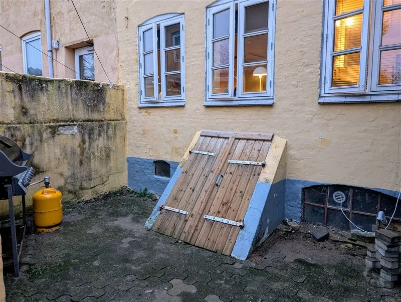 En gammel gård med en træport og gul propantank samt en mur med flere vinduer og udsigt til gården