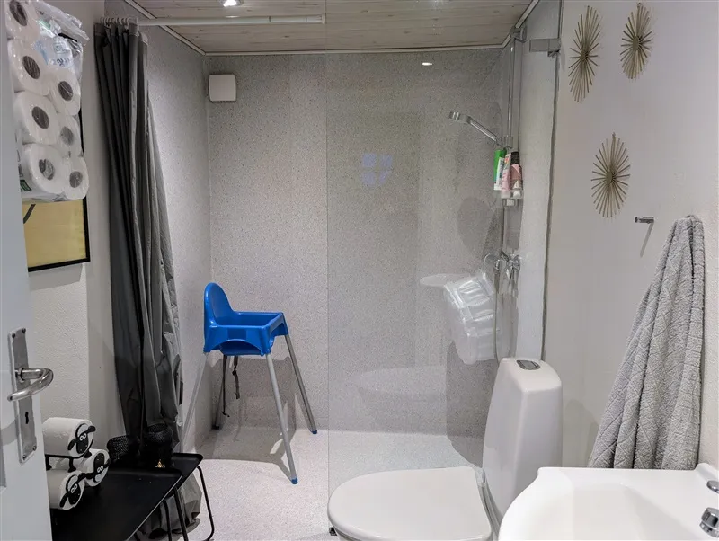 Hvidt toilet med moderne design, glasdør, og en blå barnestol. Væggene er hvide med mønstre og lysepunkter. En