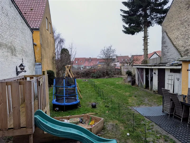 Lejlighedshaven med træskur, trampoline, legesand og græsplæne med udsigt til gavlsiderne af røde bygninger i baggrunden