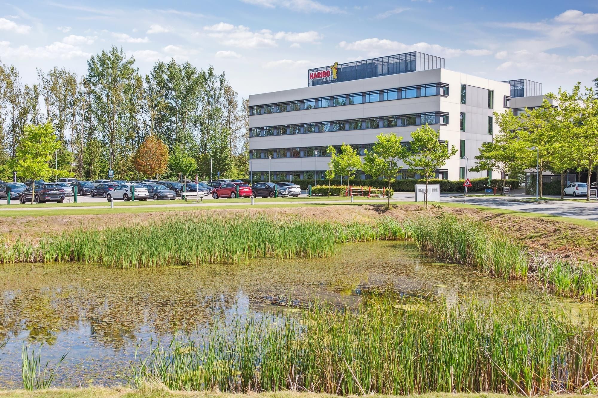 En moderne bygning med Haribo-logoet, omgivet af grønne områder, buske og sø. Parkerede biler og vejledningsskilte.