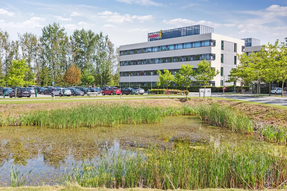 En moderne bygning med Haribo-logoet, omgivet af grønne områder, buske og sø. Parkerede biler og vejledningsskilte.