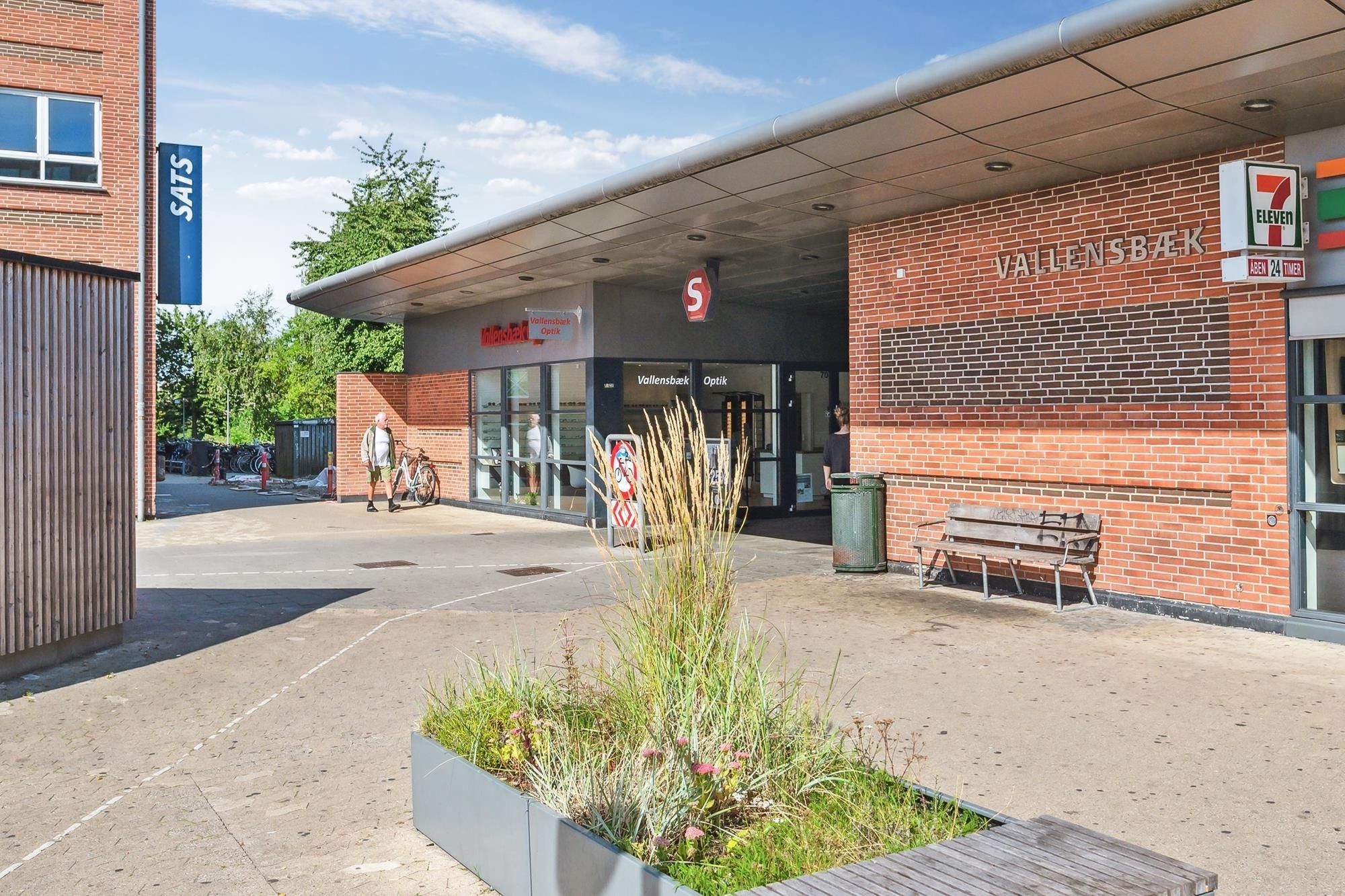 To personer går med cykler og sætter cykler ved Vallensbaek S-togstation og et bord med SATS banner