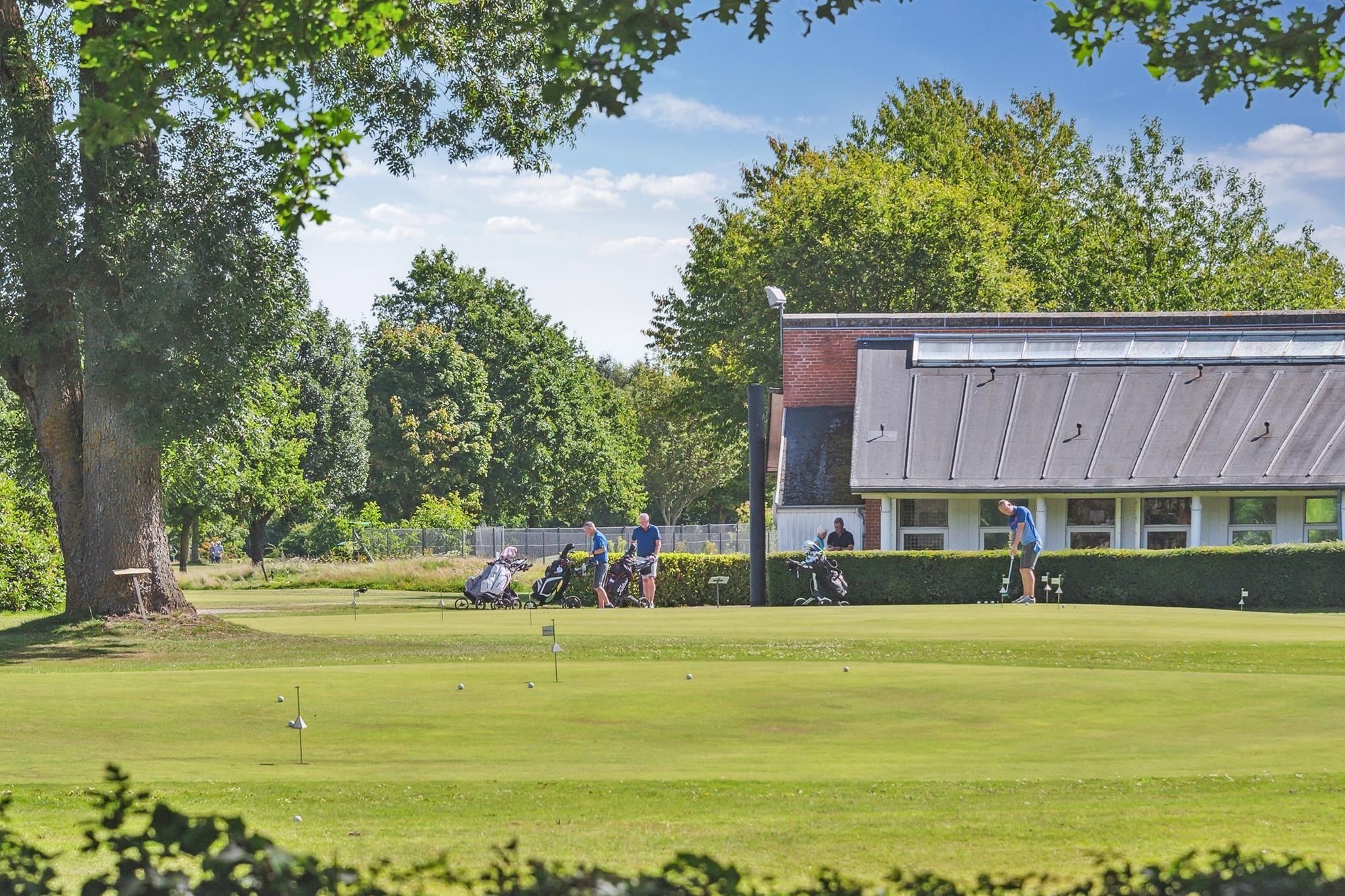 Golfere spiller golf i et lukket område på en solrig dag i sommer. Golfbanen er omgivet af grønne planter og træer. Et hus
