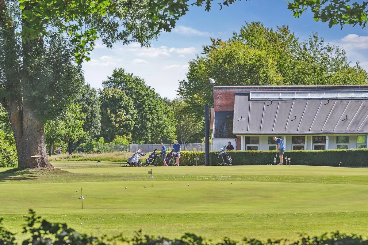Golfere spiller golf i et lukket område på en solrig dag i sommer. Golfbanen er omgivet af grønne planter og træer. Et hus