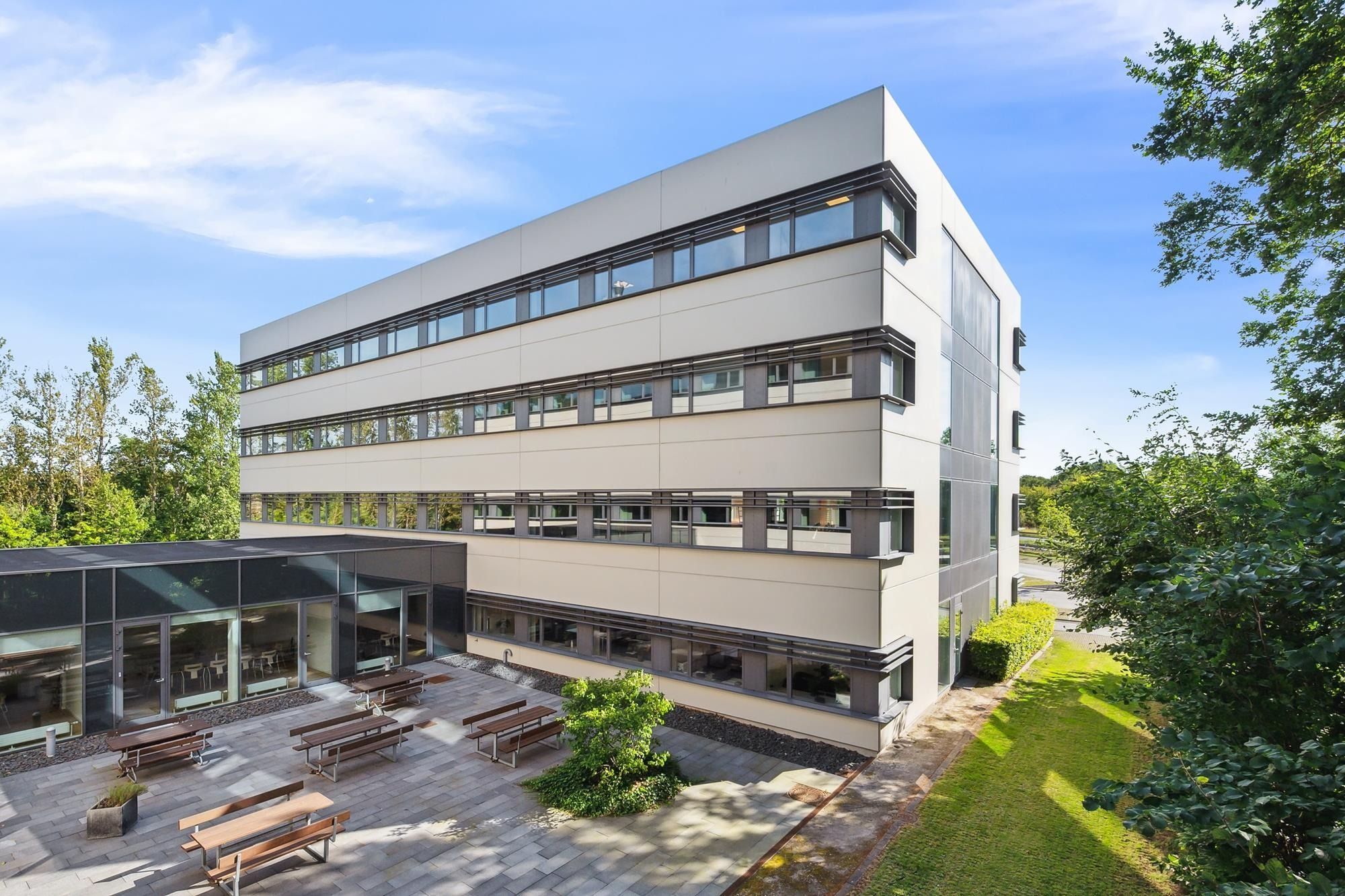 Kontorbyggeri med moderne design, glasvinduer og terrasse med bænke og plantager under en blå himmel med skyer
