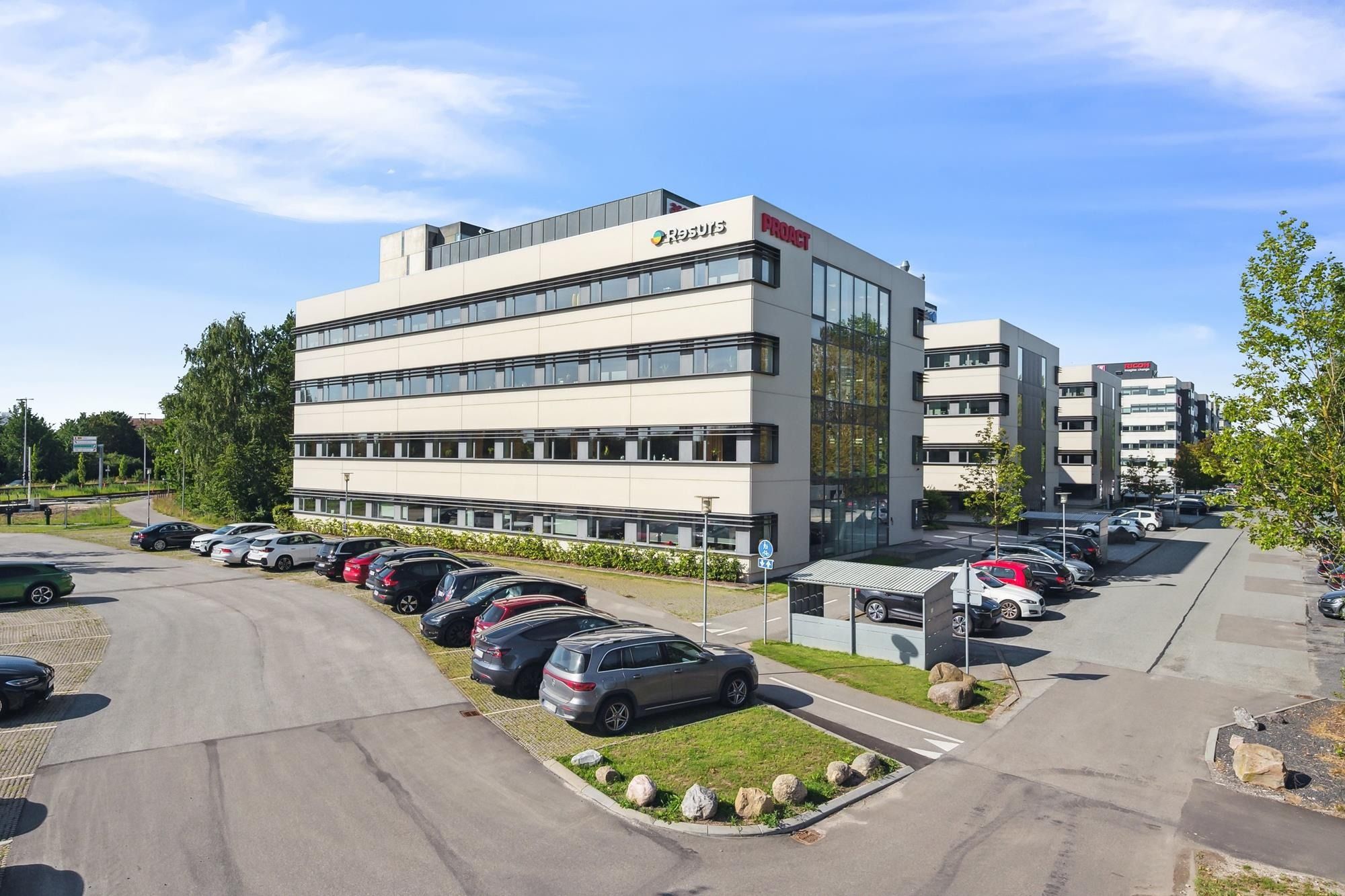 Kontorbygning med facader i lysebrune og hvide farver og logoet Resulis. Foran bygningen er der parkeringspladser og grønne