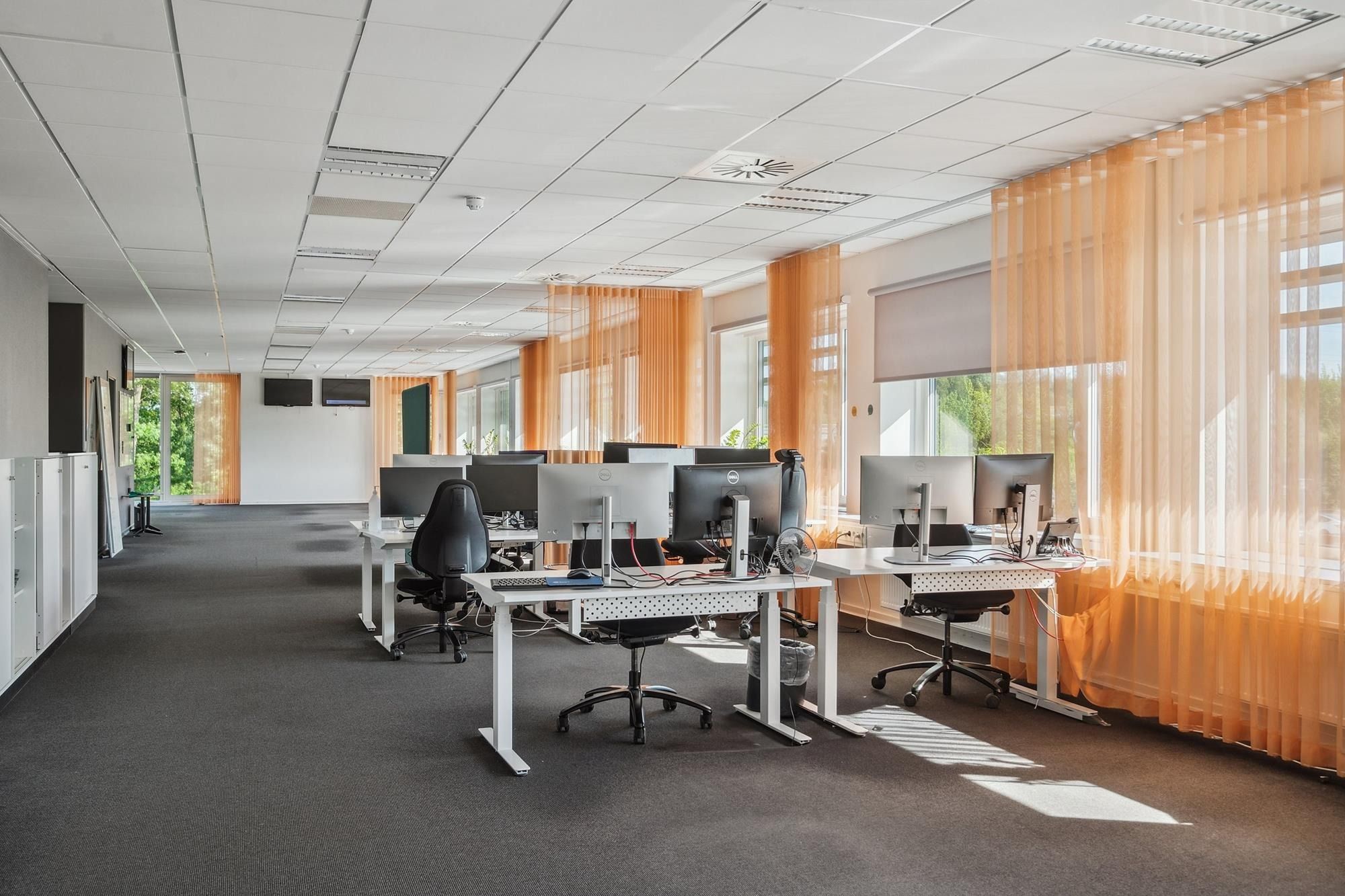 Kontor med hvide vægge, gulve og lofter. Flere bordplader med computere, skrivebordsstole og en køleskab. Vinduer med orange