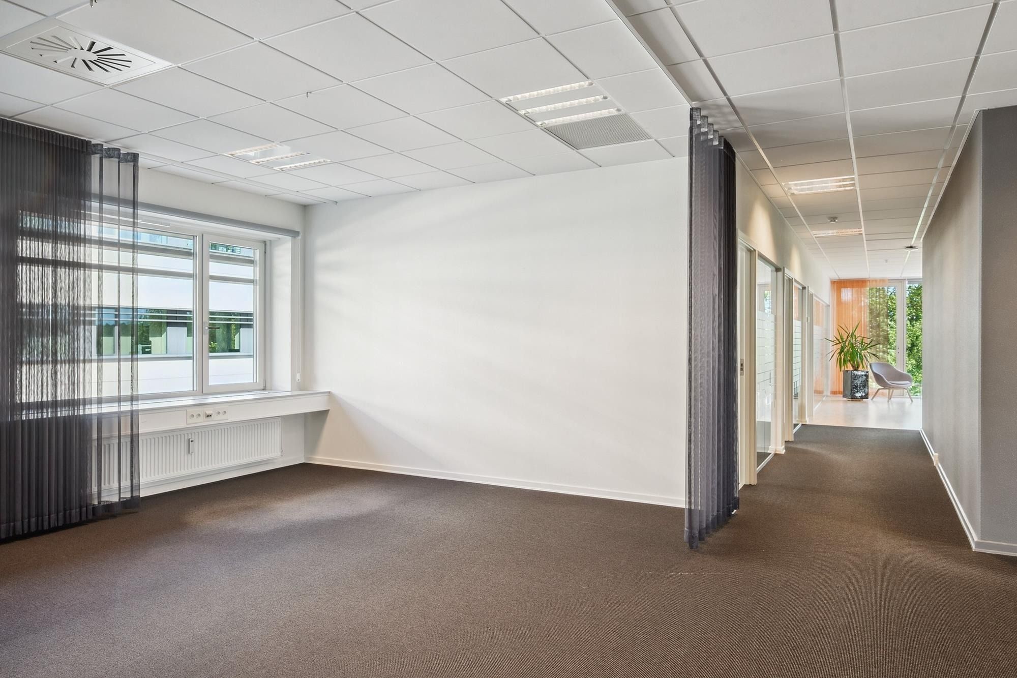 En stor, tom kontorlokal med hvide vægge og gulv, hvor der er et vindue med gardiner og et spisskur med radiatorer.