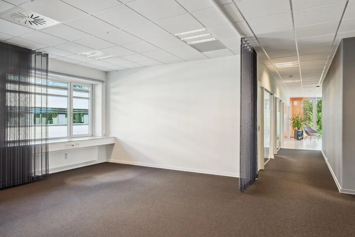 En stor, tom kontorlokal med hvide vægge og gulv, hvor der er et vindue med gardiner og et spisskur med radiatorer.