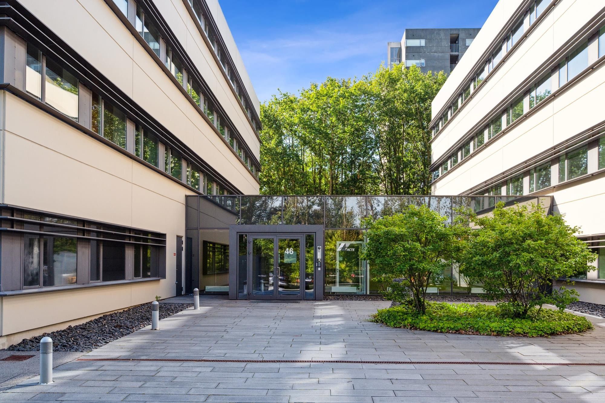 Kontorbyggeri med glasdør og vækstplanter. Solskærmet indgangszone med skriftligt FAKTE-mærke. Forskudte bollards og træer i