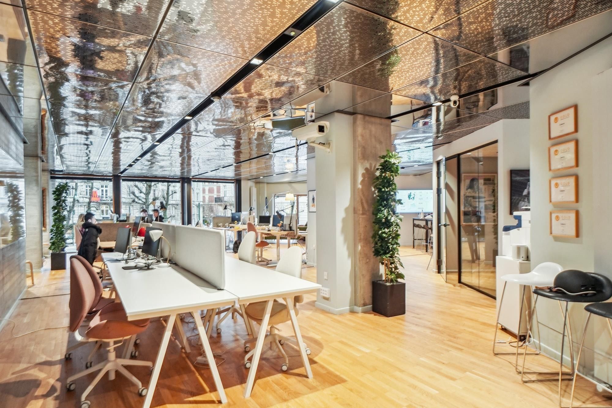 Inde i et moderne kontor med højt loft og lysegule gulve, hvor der er flere bordsæt med computere, lyse stole og høje planter