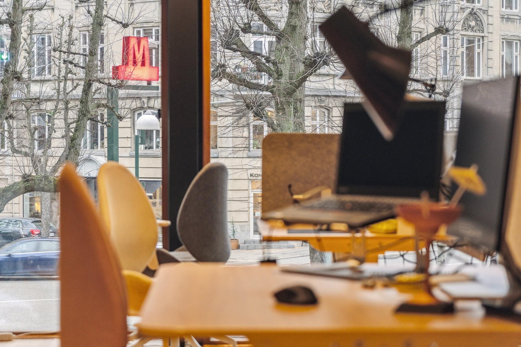 Kontorbord med monitor og mus og udsigt til gågade med biler og lysskilting