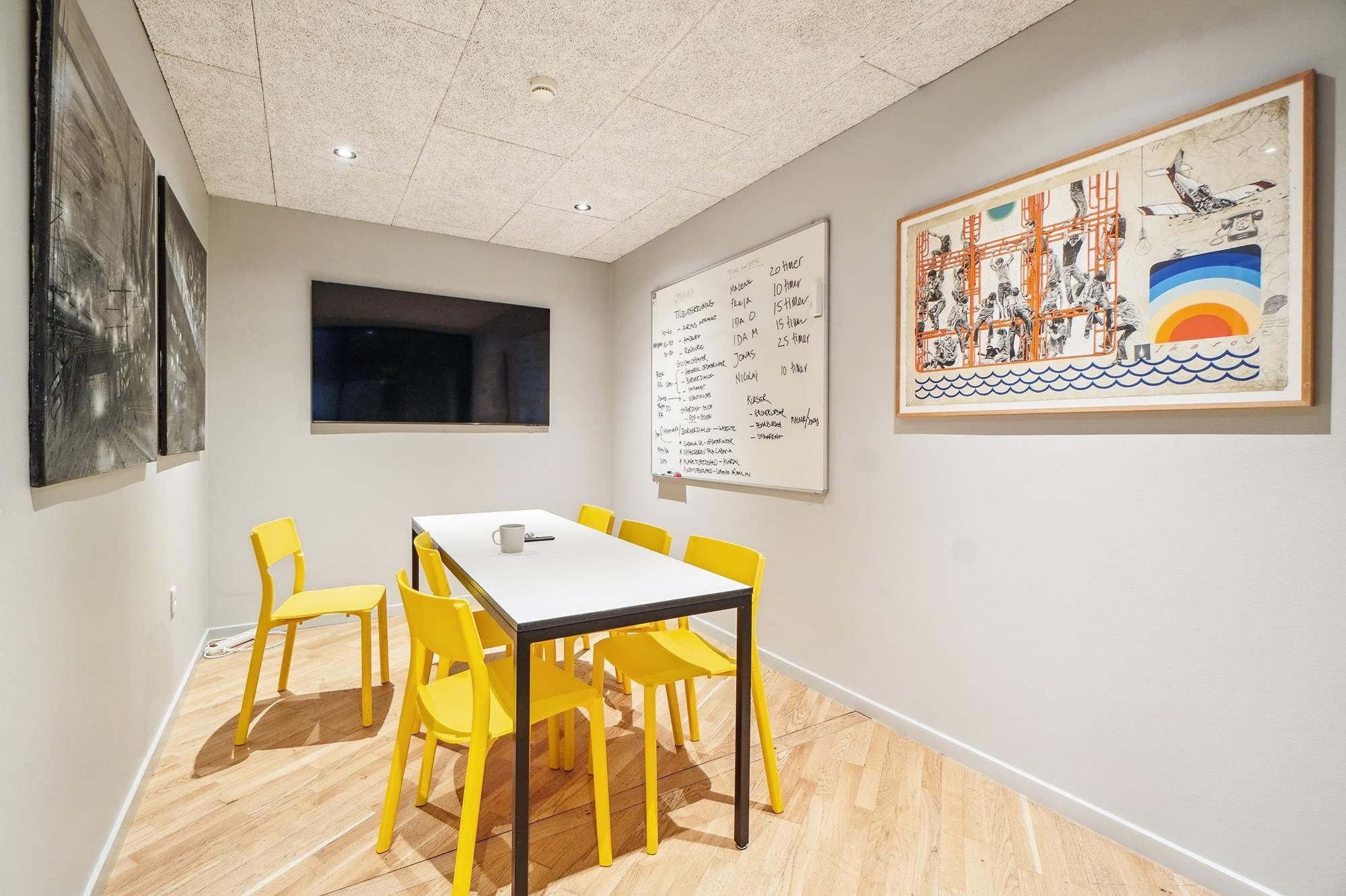 Et lille mødelokale med lysegult gulv og hvide vægge. På bordet er der en kop og en fjernbetjening. Væggen har en