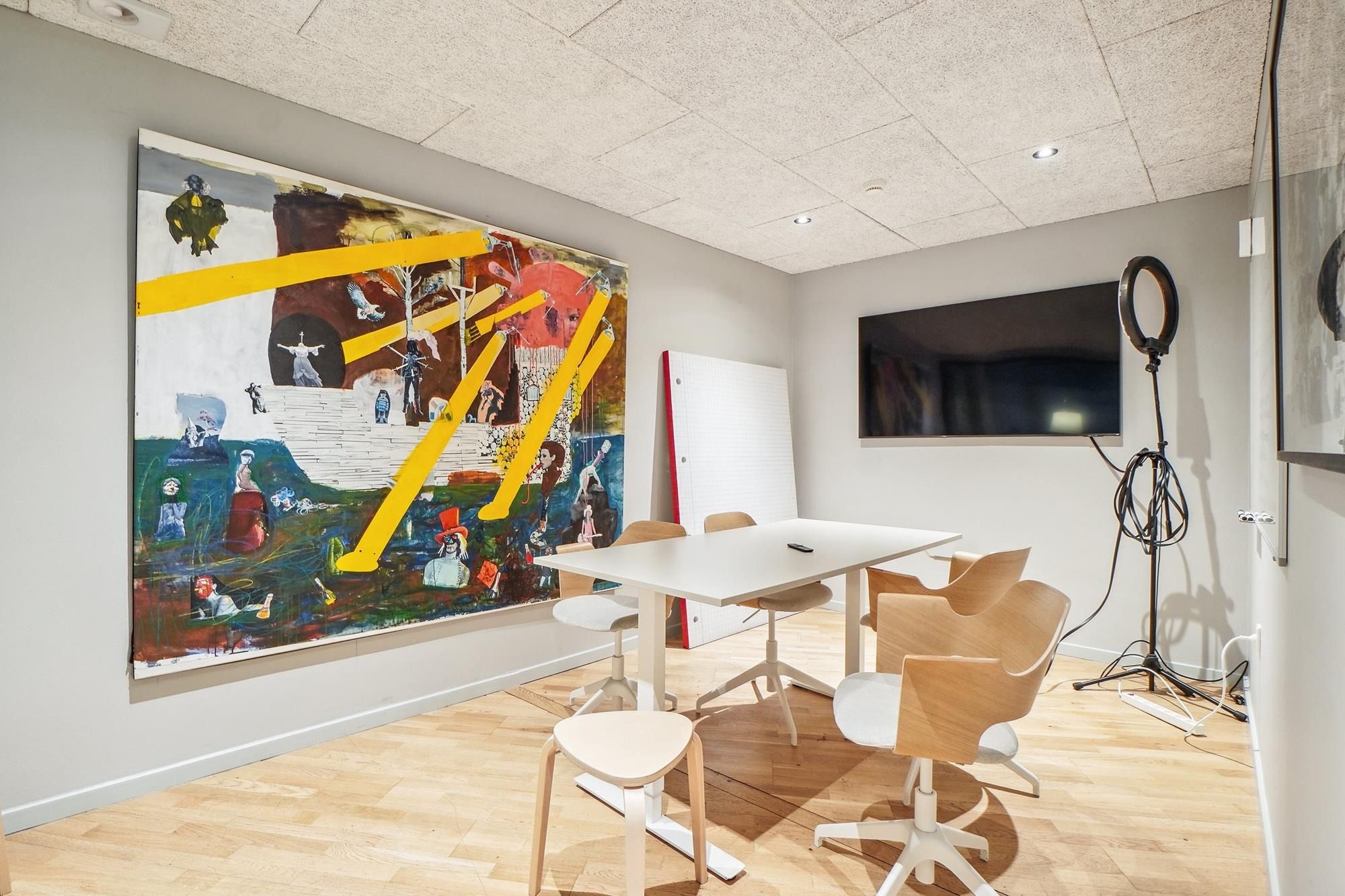 I et kontorlokale er der en stor maleri på væggen, et hvidt rundt bord og flere stole, samt en stor skærm på væggen og en
