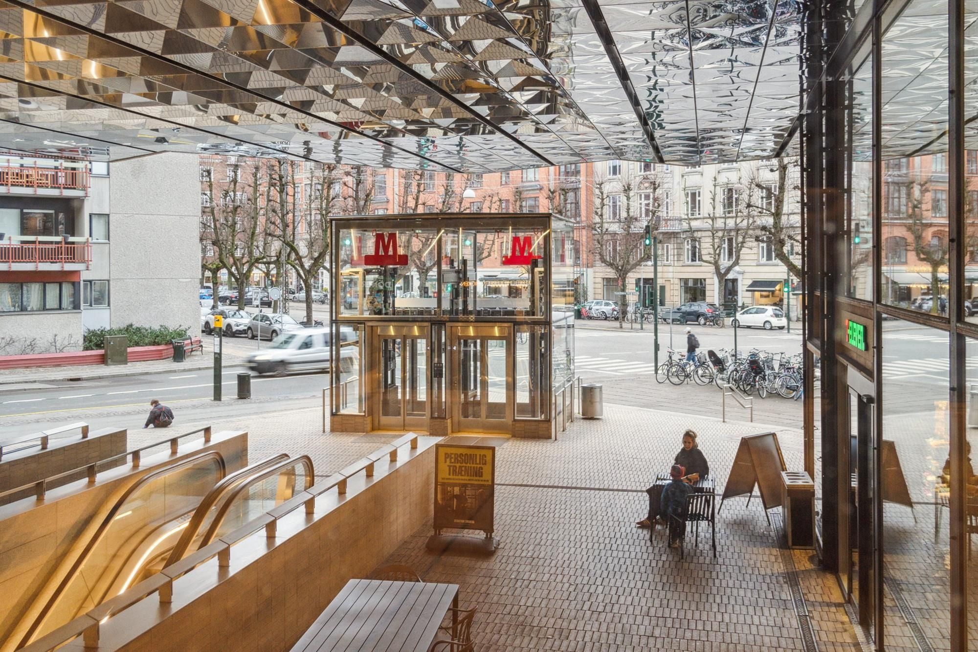 To mennesker sidder på en cafeterrasse udenfor med bord og stole og flere mennesker er på cykler og biler på vejen