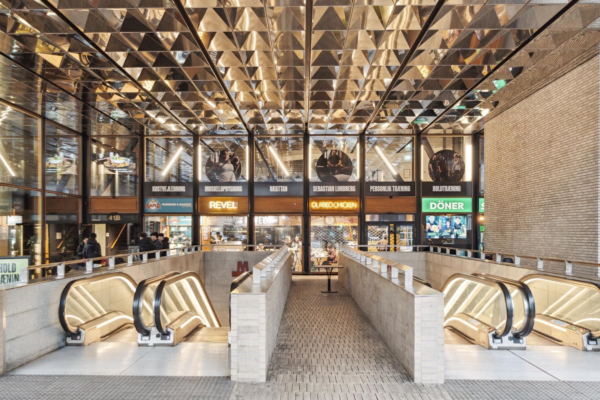 Tunnelbanestation med opgange og store vinduer i højre side. Restauranter med navnene Sebastian Lundberg, DLFRIEDCHICKEN og