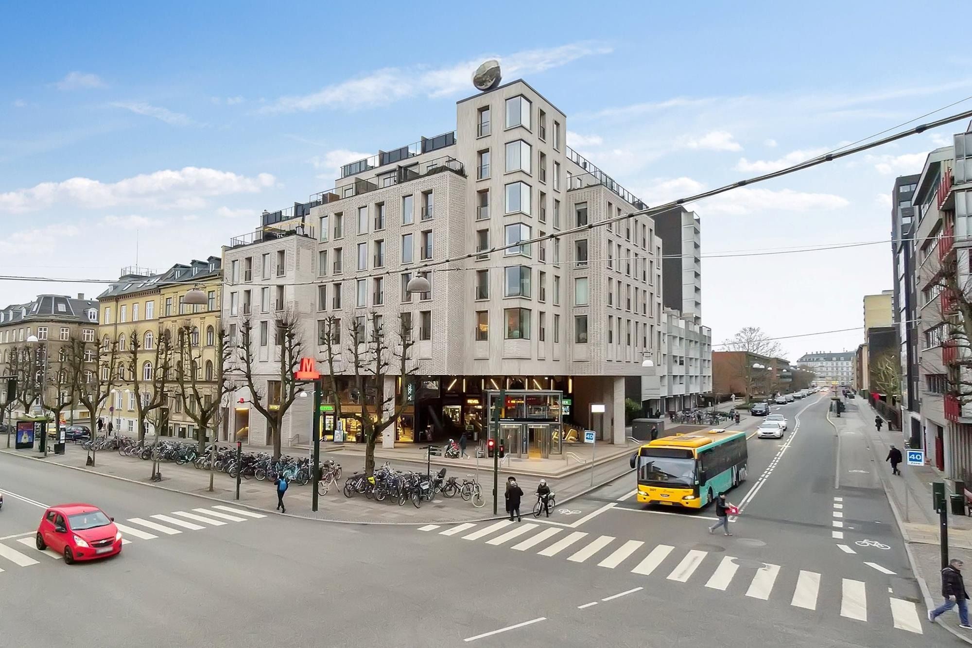 Frederiksberg Allé med en gågade, rødklappet bil og gul bus samt flere fodgængere, et stort hvidt bygningskompleks med en