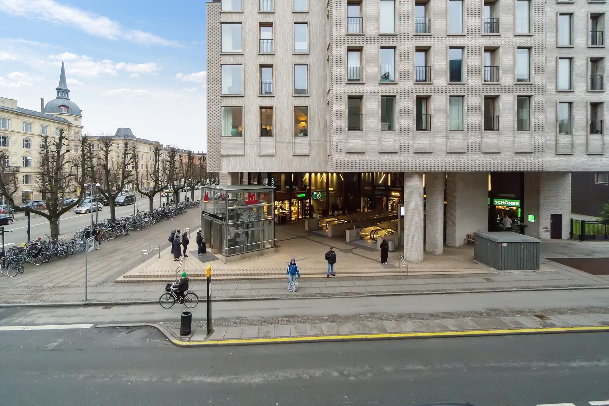 Folk går på gågaden, mens en cykelkører kører forbi. På hjørnet er der et hus med mange vinduer, et stort indkøbscenter og