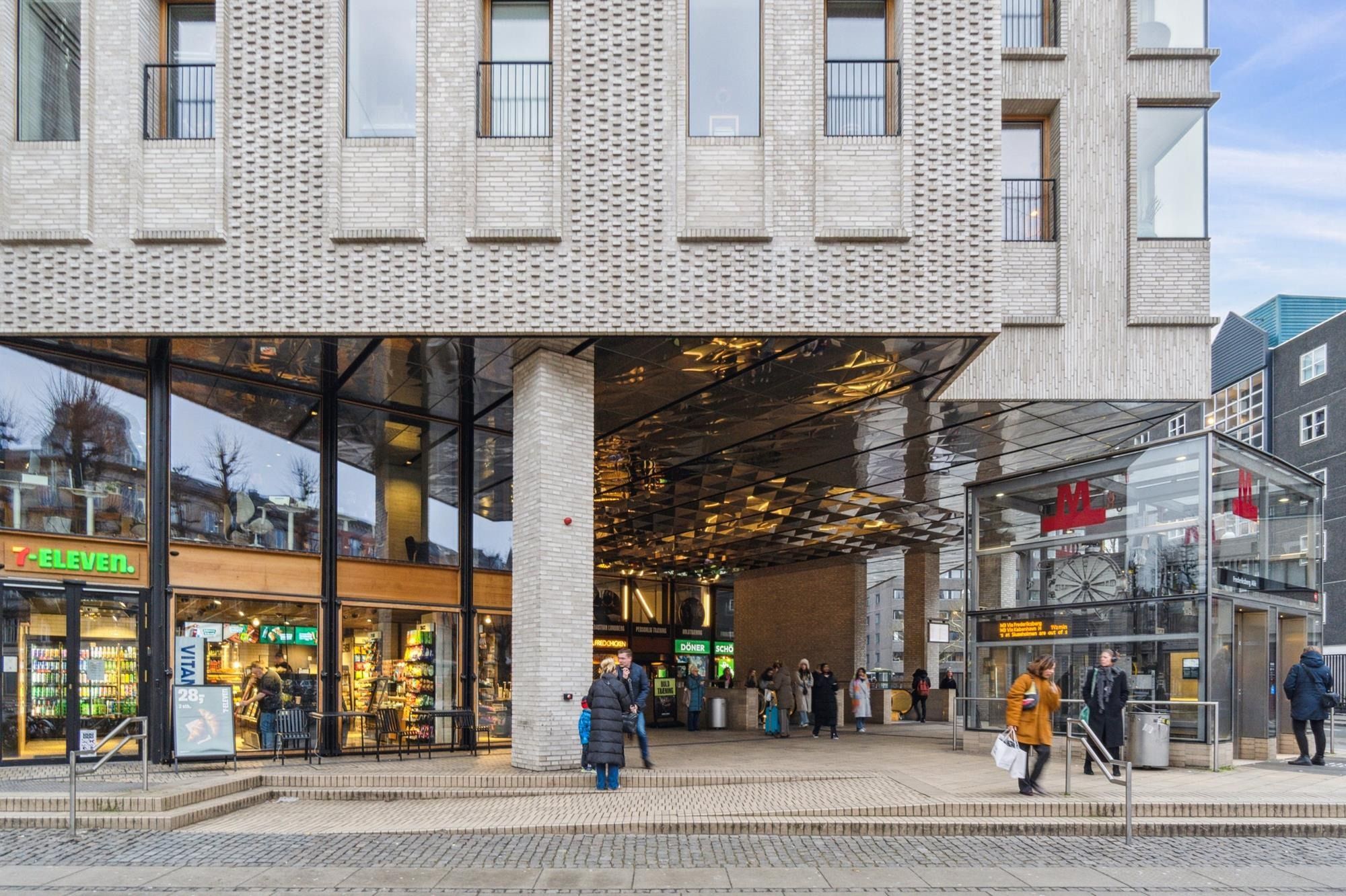 En gruppe mennesker gå forbi en butik med 7-Eleven-skilt og et salgsskilt for Vitan-produkter, samt en kiosk med et stort