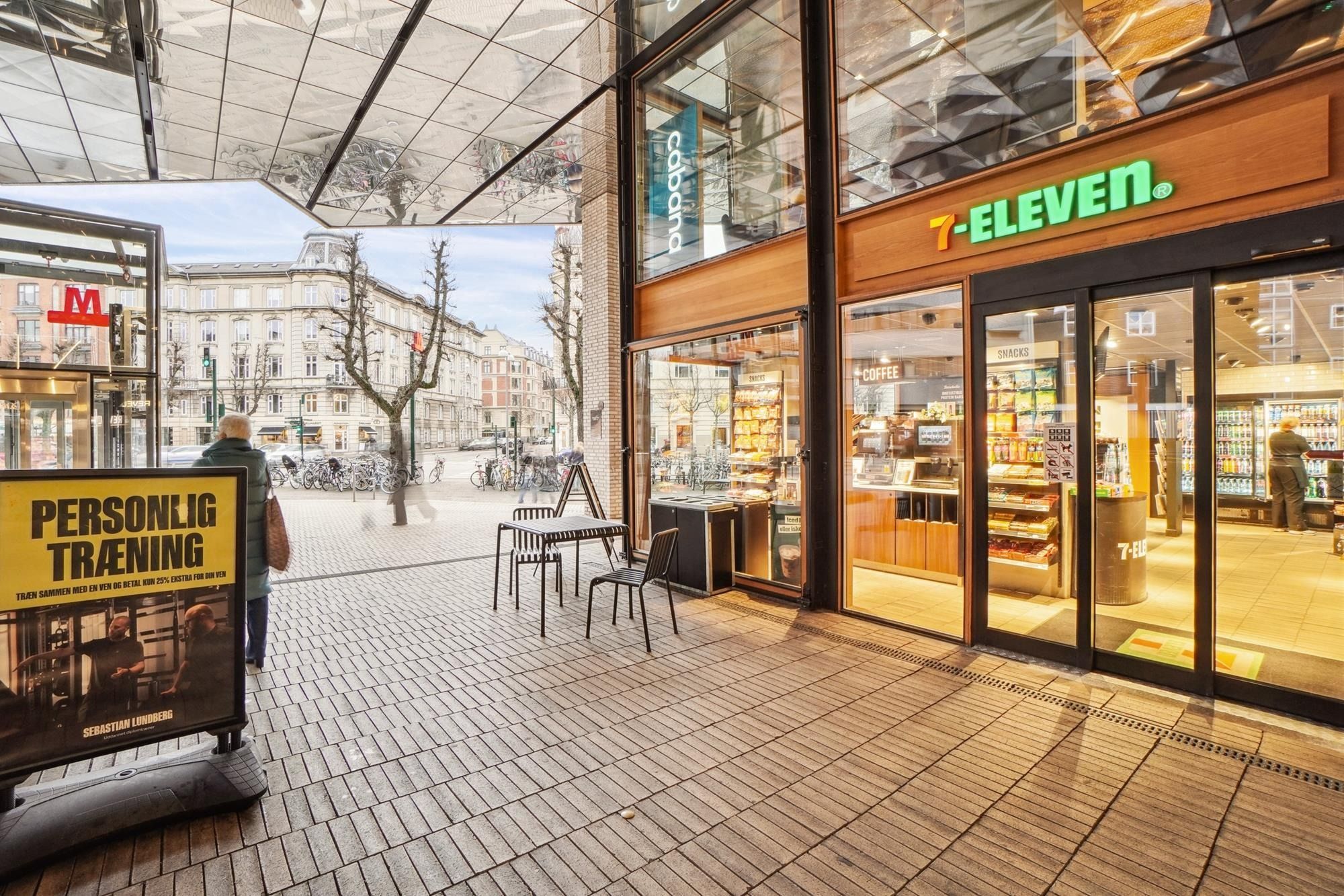 På en torgplads står en skilt med teksten "Personlig træning" og en reklame for Sebastian Lundberg. Der er en 7-Eleven-butik