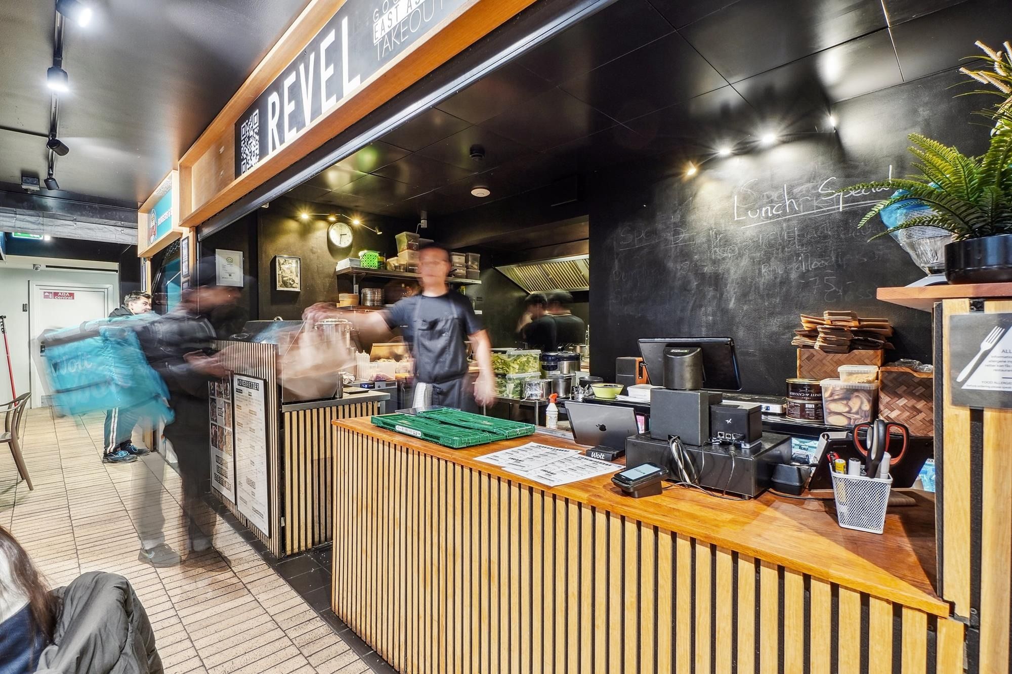 Butik med rødt REVEIL-logo på facaden, en kassaskranke og grønne plantekrukker med papirposer og produkter, en sort