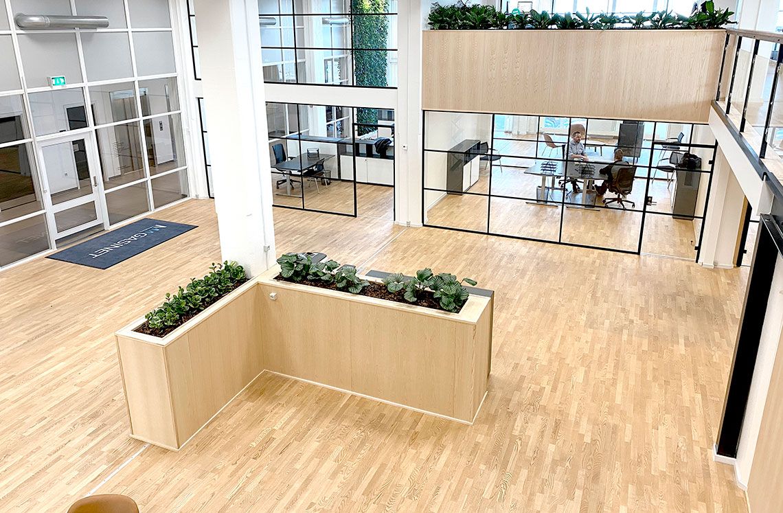 Kontorlokaler med store glasruder og trægulve, kontormøbler, plantepot, sofa, mennesker og skrivebord.