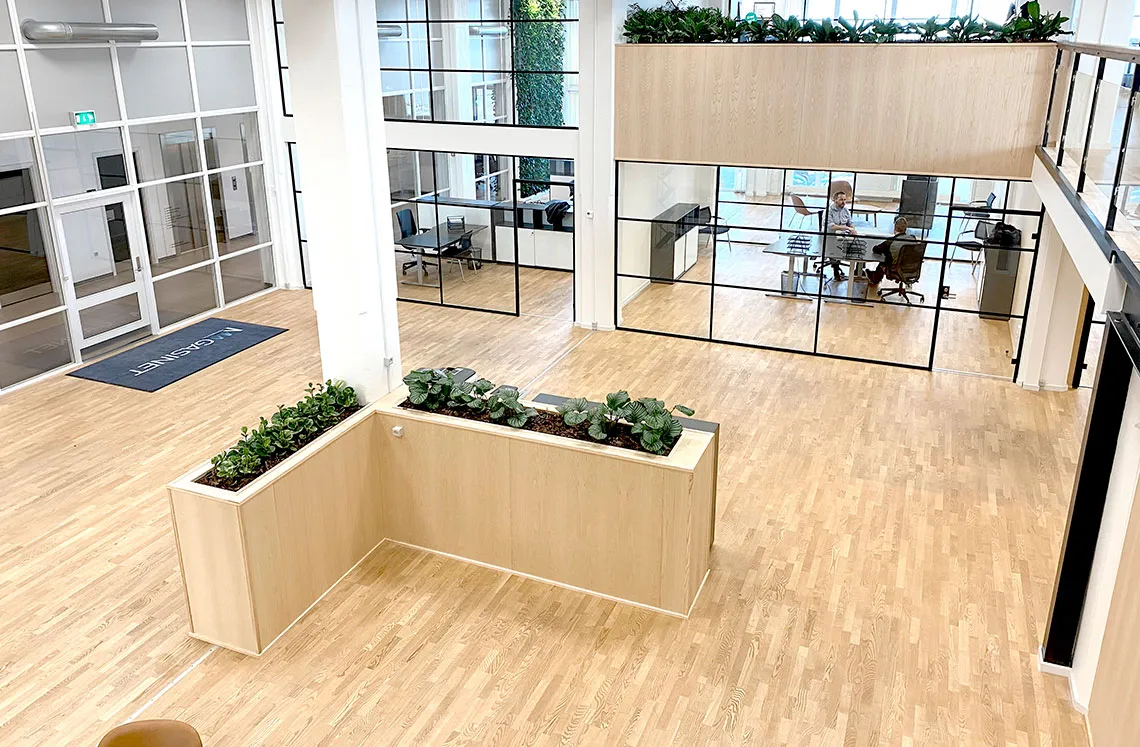 Kontorlokaler med store glasruder og trægulve, kontormøbler, plantepot, sofa, mennesker og skrivebord.