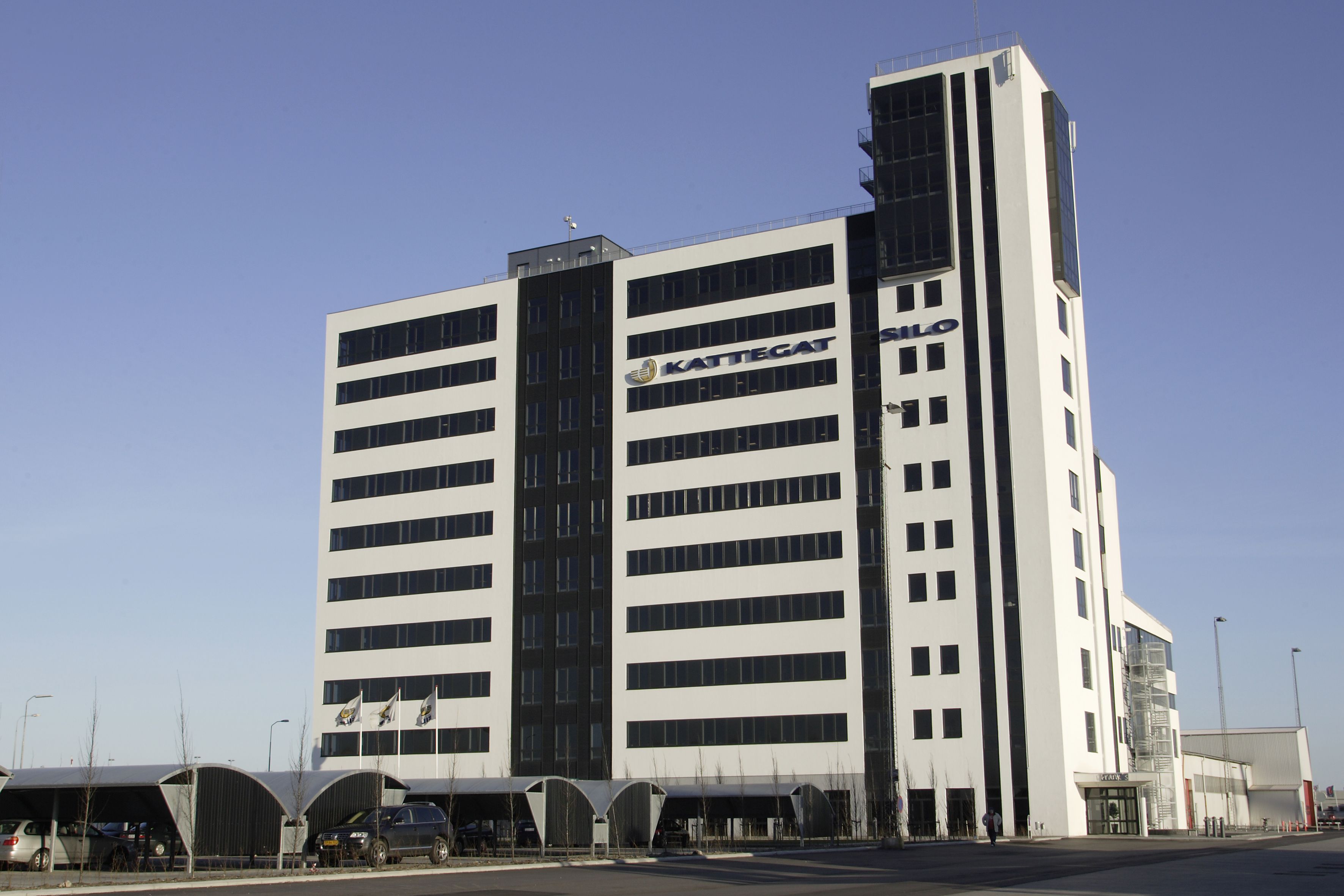 Et højt hvidt og sort bygning med KATTEGAT-logoet på siden og flere flag samt biler parkeret i skyggede parkeringspladser