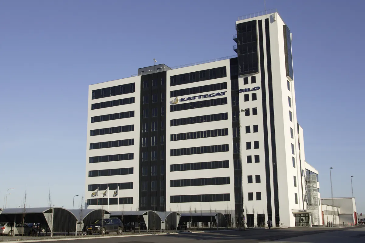 Et højt hvidt og sort bygning med KATTEGAT-logoet på siden og flere flag samt biler parkeret i skyggede parkeringspladser