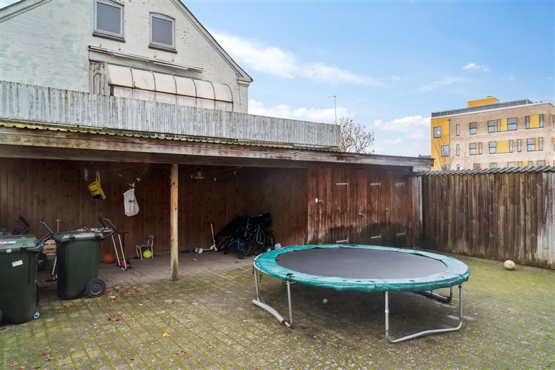 En gade med et hus og garage med trampoline, affaldsposer og cykler samt en bygning med gul facade