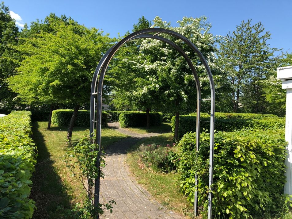 En bueformet gangbro i en velplejet have med buske, græs og træer i baggrunden
