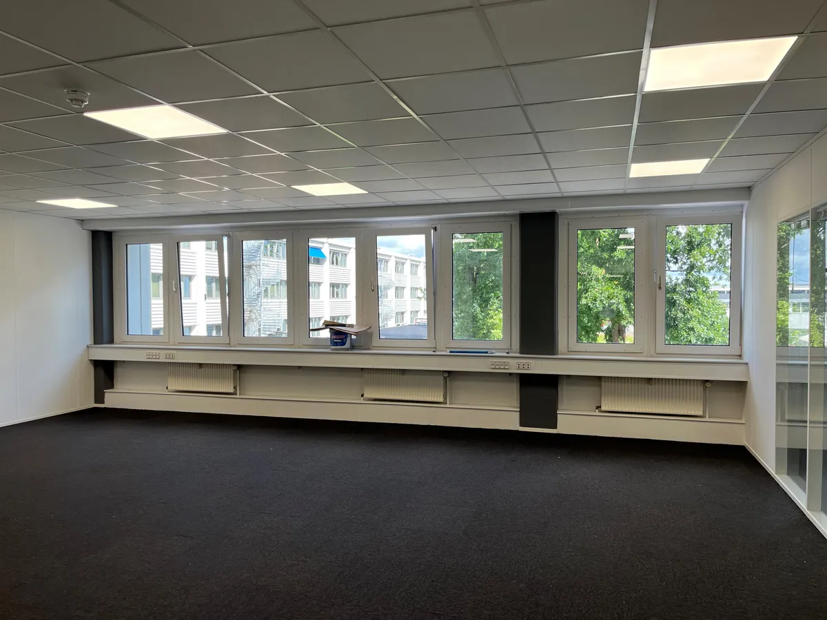 Store glasruder med udsigt til bygninger og grønne træer samt hvide vægge, mørke gulvbelægning og hvide loftsplader med