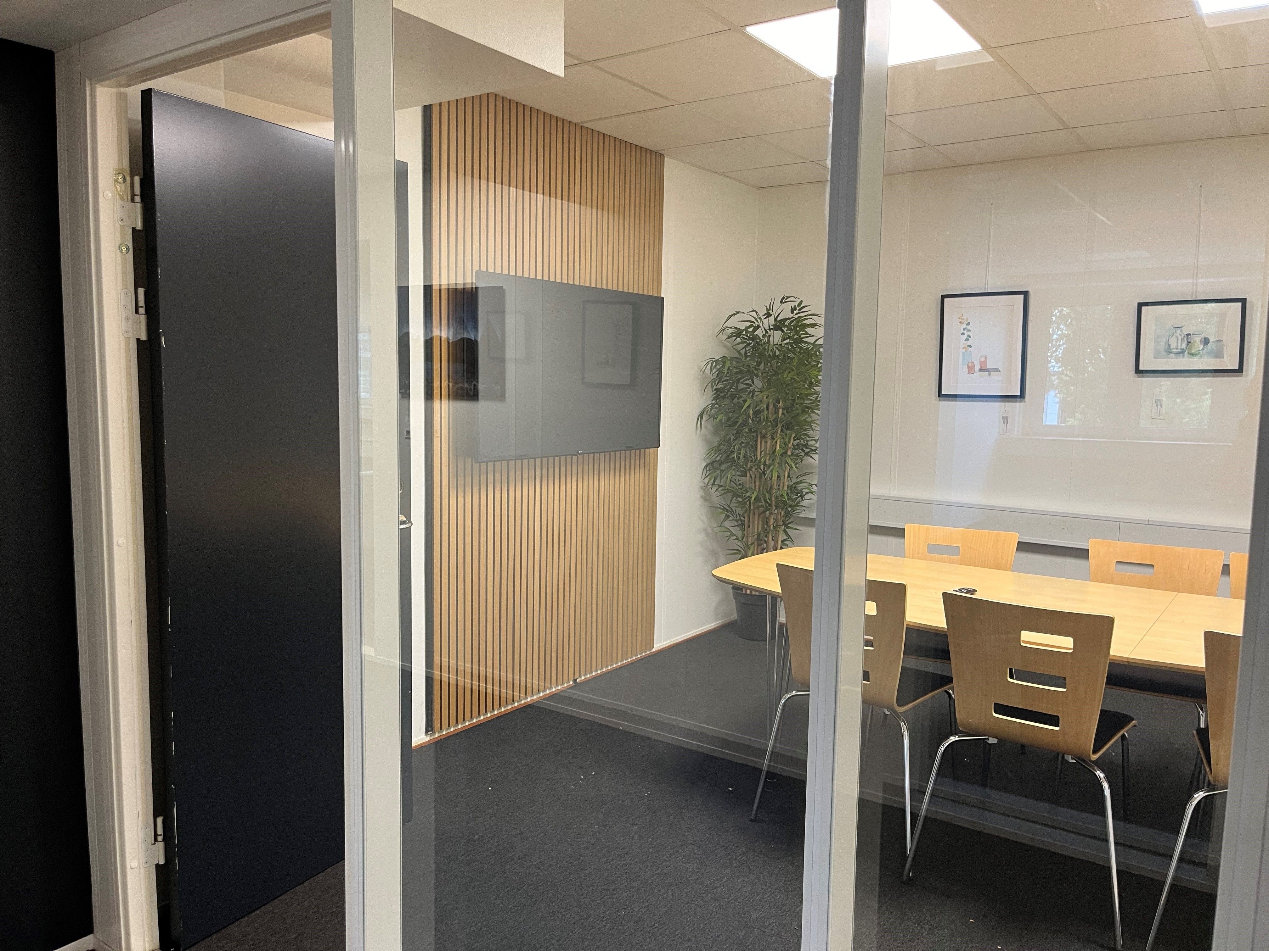 Moderne konferencerum med sort dør og lysegul gulvbelægning og tre træstole med ryglæn under langt, brunligt bord