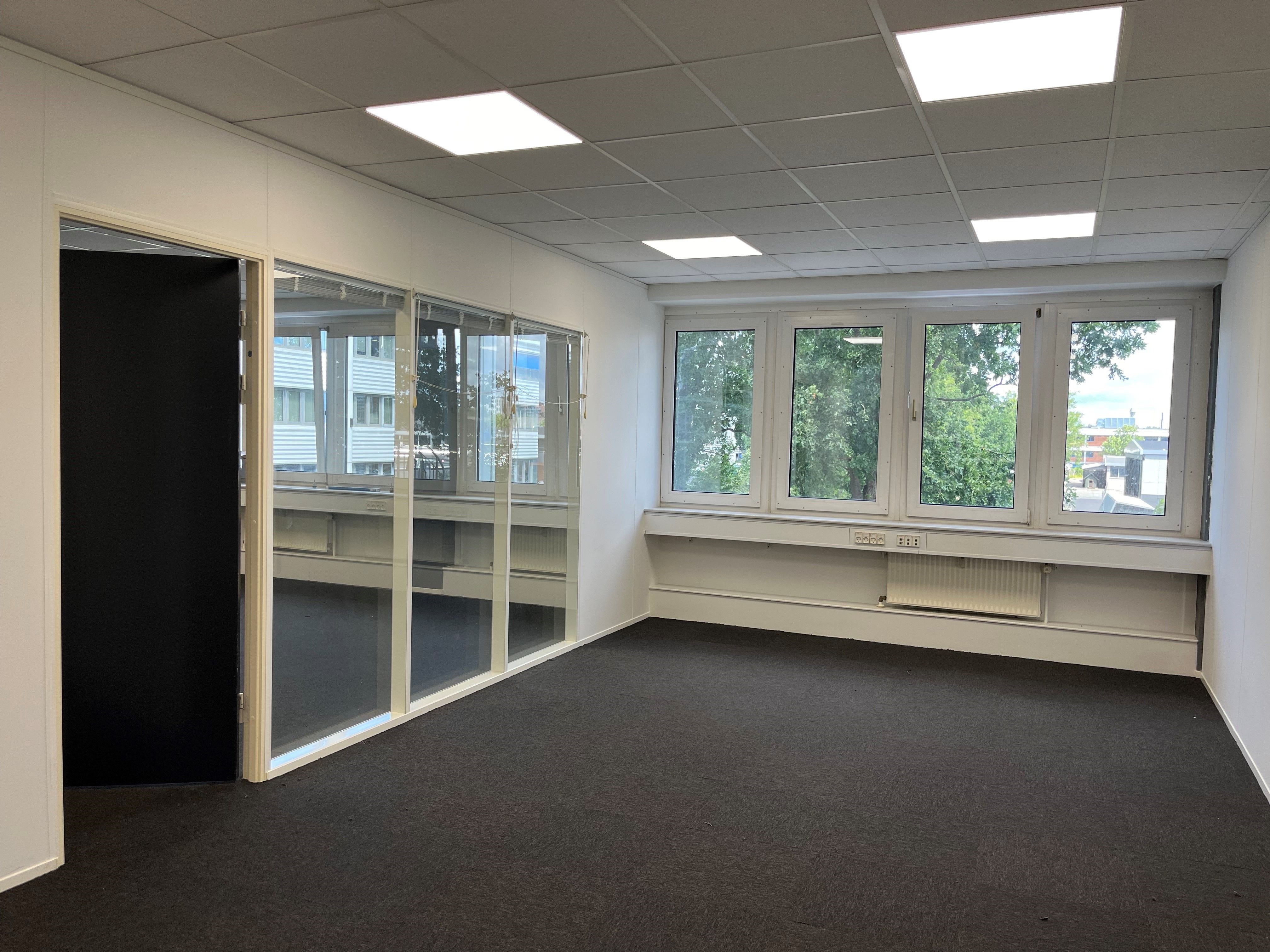 Tomt kontor med lysegul væg, sort gulv og store vinduer med udsigt til bygninger og træer.