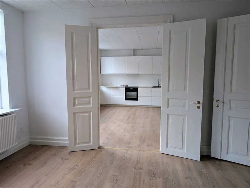 To hvide døre står åbne og viser et lyse hus med køkken med hvide skabe og en sort indbygget ovn