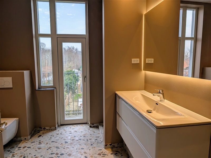 Badrum med toilet, vask, spejl og lyspærer. Et lyse, moderne badrum med lysegul væg, lysgrå gulv og hvidt toilet.